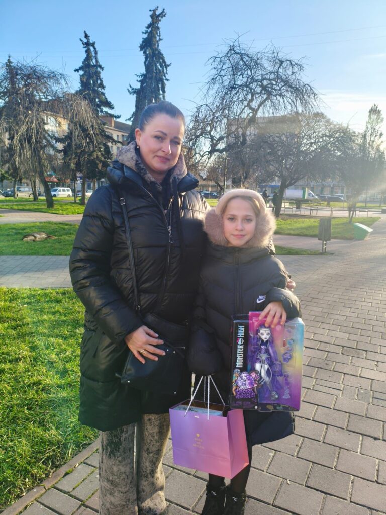 Двоє людей стоять на мощеній доріжці в парку. Обоє — у чорних зимових куртках. Один тримає коробку з лялькою «Monster High» і фіолетовий пакет із написом «SWEET MAGIC». Позаду — зелена трава та голі дерева, день сонячний. Сцена — зимова, момент вручення подарунка