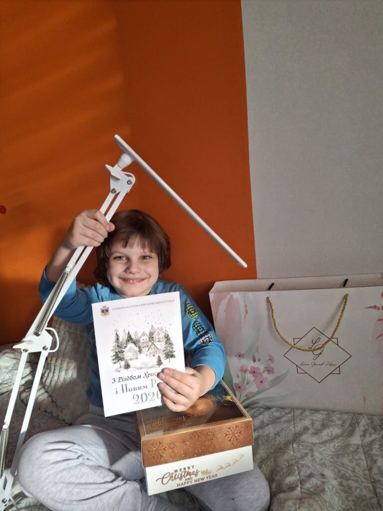 Дитина сидить на ліжку, усміхається, тримає білу настільну лампу та листівку з зимовим пейзажем. Перед дитиною — коробка з написом «Merry Christmas and Happy New Year» і подарунковий пакет із квітковим дизайном та написом «Luxe Grand Place».