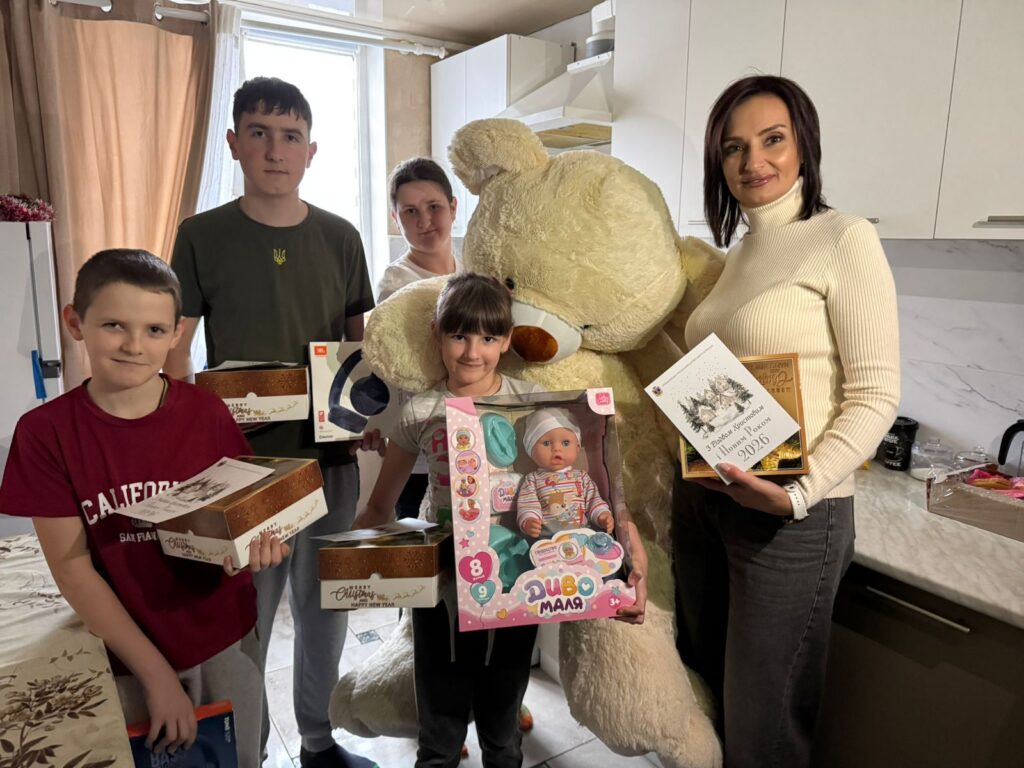 У кухні стоять п’ятеро людей із подарунками. Видно великого плюшевого ведмедя, коробку з написом «Happy New Year», ляльку «Диво Маля», навушники та листівку з привітанням «З Новим Роком та Різдвом Христовим! 2024». Інтер’єр — сучасний, із білими шафами та мармуровою стільницею. Атмосфера — святкова, тепла.