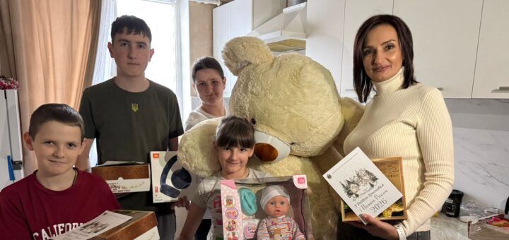 У кухні стоять п’ятеро людей із подарунками. Видно великого плюшевого ведмедя, коробку з написом «Happy New Year», ляльку «Диво Маля», навушники та листівку з привітанням «З Новим Роком та Різдвом Христовим! 2024». Інтер’єр — сучасний, із білими шафами та мармуровою стільницею. Атмосфера — святкова, тепла.