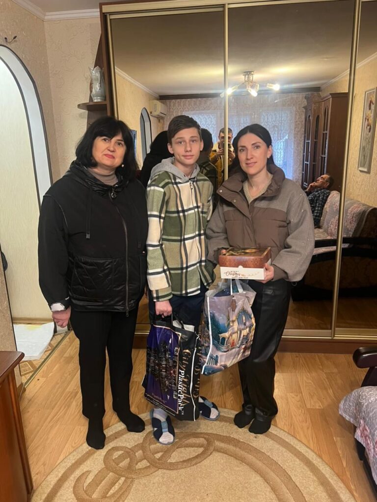 У кімнаті з дзеркальною шафою стоять троє людей. Посередині — хлопчик у зеленій картатій толстовці з подарунковим пакетом «Philadelphia». Праворуч — людина з коробкою та святковим пакетом. У дзеркалі видно ще двох людей, один із них фотографує. Інтер’єр — теплий, із меблями, картиною та фігуркою на полиці. Сцена — домашня, з елементами святкування.