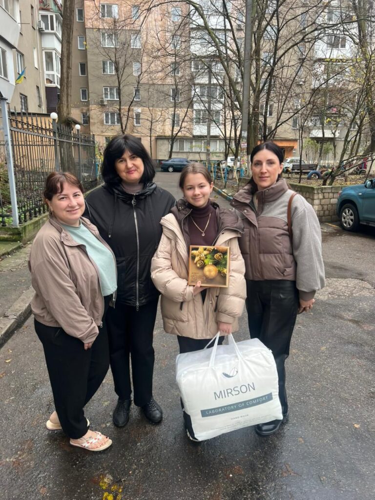 Четверо людей стоять на вулиці перед багатоповерховим житловим будинком. У центрі — дівчинка з подарунковою коробкою з написом «2024 З Новим Роком!» і великою білою сумкою з написом «MIRSON LABORATORY OF COMFORT HAND MADE». Усі вдягнені тепло, на фоні — голі дерева. Атмосфера — зимова, момент вручення подарунків.