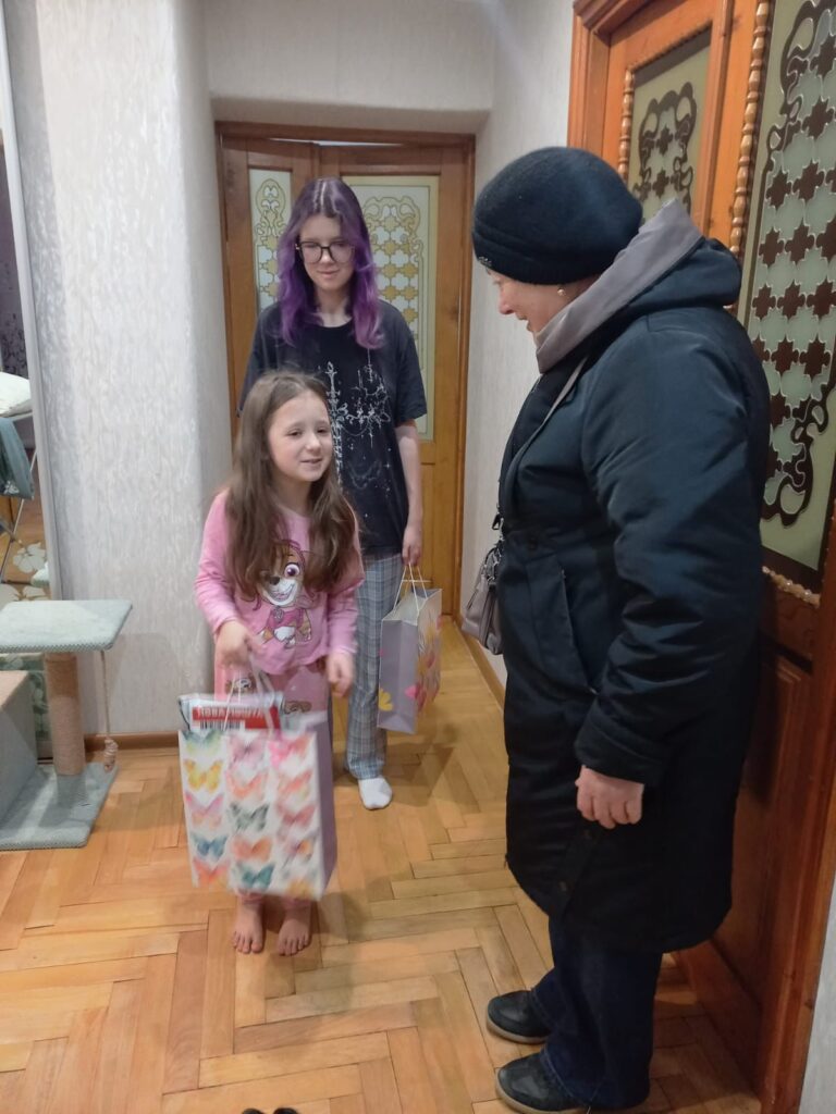 У домашньому коридорі стоять троє людей. Маленька дитина в рожевій піжамі тримає яскравий подарунковий пакет із метеликами. Поруч — старша людина в чорному пальті та шапці, а позаду — доросла з фіолетовим волоссям у картатих піжамних штанах. Видно дерев’яну підлогу, декоративні двері з матовим склом і когтеточку для кота.