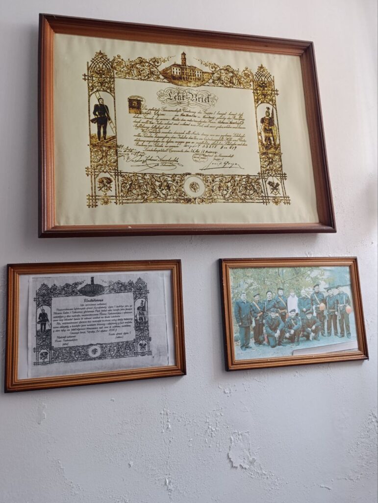 На білій стіні — три підвісні рамки. Верхня — кольоровий сертифікат із золотими орнаментами, фігурами в традиційному вбранні та будівлею вгорі; текст — німецькою, про завершення навчання. Ліворуч унизу — чорно-білий сертифікат із подібним оформленням і німецьким текстом. Праворуч унизу — кольорова фотографія групи людей у формі, ймовірно історичне або церемоніальне фото.