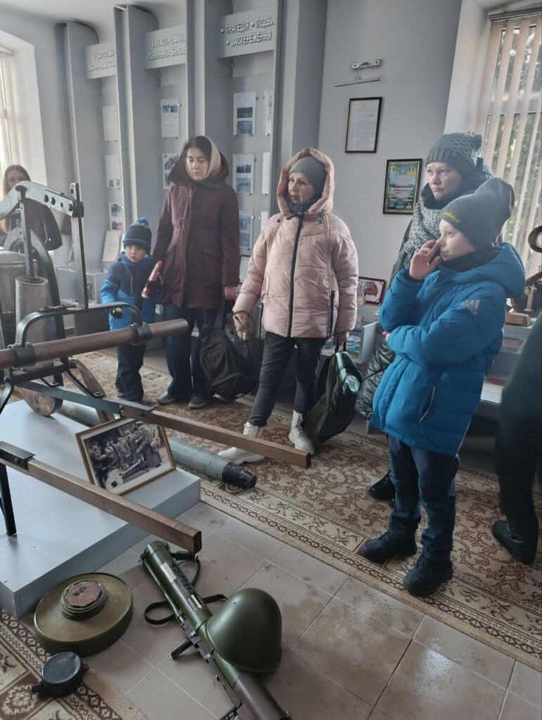 У музейному просторі група дорослих і дітей розглядає експозицію з військовими артефактами: гранатомет, зелений шолом, міна, чорно-біле фото солдатів. Позаду — вертикальні панелі з українськими заголовками: «Героїзм воїнів-захисників», «Катастрофи, пожежі на блоков», «Трагедія — подвиг — застереження».