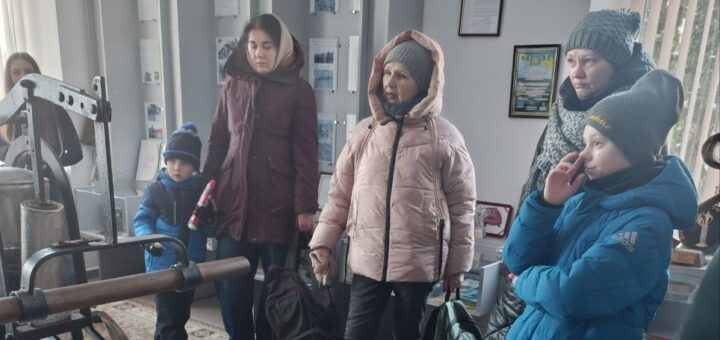 У музейному просторі група дорослих і дітей розглядає експозицію з військовими артефактами: гранатомет, зелений шолом, міна, чорно-біле фото солдатів. Позаду — вертикальні панелі з українськими заголовками: «Героїзм воїнів-захисників», «Катастрофи, пожежі на блоков», «Трагедія — подвиг — застереження».