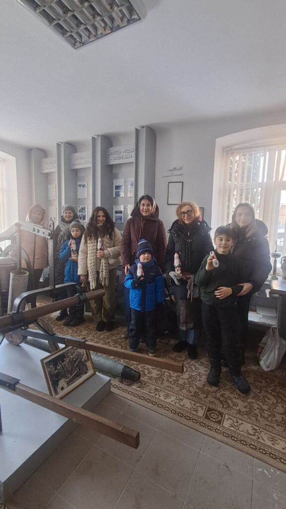 У виставковій кімнаті група дорослих і дітей позує на фоні експозиції зі старим обладнанням. Декілька людей тримають фігурки Діда Мороза, що натякає на святковий контекст. Позаду — панелі з текстами кирилицею, фотографіями й документами. На постаменті — чорно-біле фото.