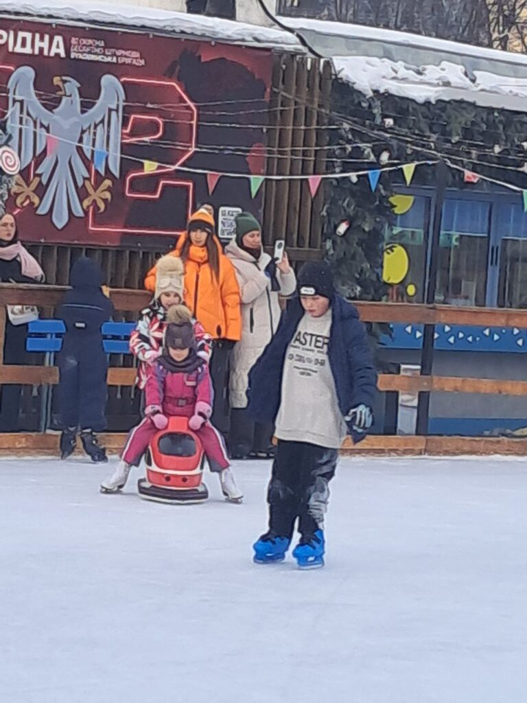 На ковзанці — дитина в синіх ковзанах і чорному одязі з написом «MASTER» на світшоті. Позаду — дитина в рожевому комбінезоні сидить на червоній фігурці-помічнику у формі машинки, її штовхає інша дитина в яскравій куртці. За огорожею — дорослі з телефонами. На фоні — стіна з орлом, літерою «Р» і написами «РІДНА», «Це організація козацького братства Українська Рідна».