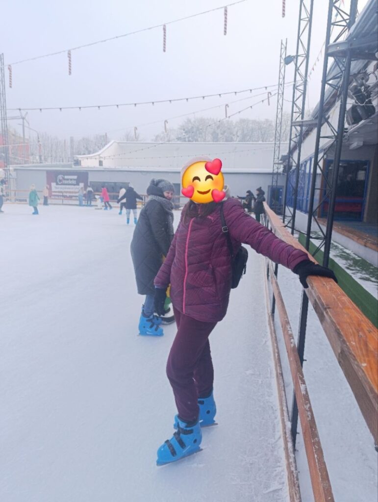 На передньому плані — людина в рожевій шапці та бордовому одязі тримається за дерев’яну огорожу ковзанки. Поруч — ще один учасник у темному пальті. Позаду — інші ковзанярі, будівля з вивісками, засніжені дерева та гірлянди над льодом.