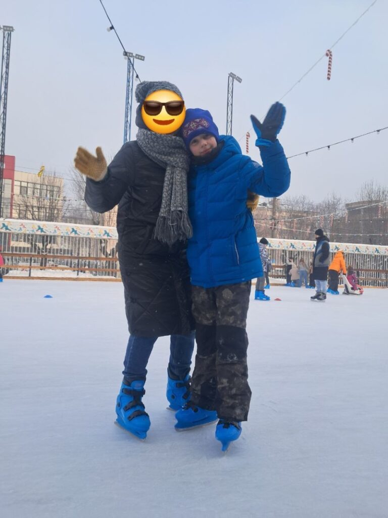 Двоє людей стоять на ковзанці, усміхаються й махають у камеру. Обидва — в синіх ковзанах і зимовому одязі. Один — у чорному пальті з сірим шарфом, інший — у синій куртці, камуфляжних штанах і шапці з червоно-жовтим візерунком. Позаду — ковзанярі, прикраси у вигляді цукерок, міські будівлі.