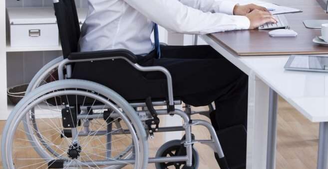 У сучасному офісному просторі людина в інвалідному візку працює за комп’ютером. Одяг — біла сорочка та темні штани. На столі — чашка, канцелярія, організовані полиці. Візок розташований близько до столу, поза — активна, зосереджена.