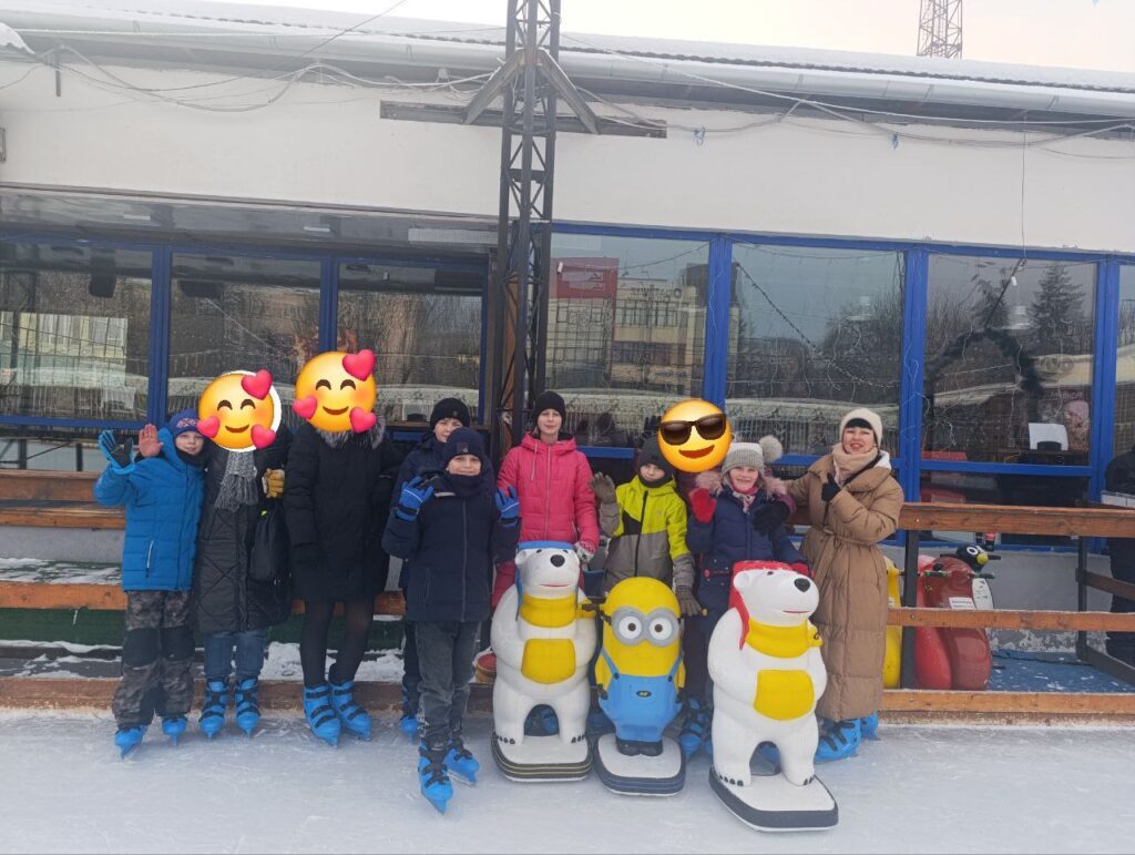 Група дітей і дорослих позує на відкритій ковзанці перед будівлею з великими синіми вікнами. Усі — в ковзанах і зимовому одязі. Попереду — три фігурки-помічники для катання: два білих ведмедики та Міньйон.