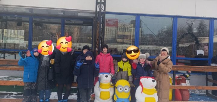 Група дітей і дорослих позує на відкритій ковзанці перед будівлею з великими синіми вікнами. Усі — в ковзанах і зимовому одязі. Попереду — три фігурки-помічники для катання: два білих ведмедики та Міньйон.