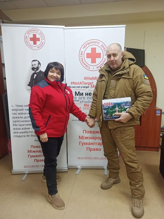 У приміщенні стоять двоє людей перед двома вертикальними банерами Червоного Хреста. Один — у червоно-чорній куртці з емблемою організації, другий — у військовій формі, тримає календар із написом «2026». Вони тримаються за руки й усміхаються. Банери містять емблеми, цитати українською про захист мирного населення, заклики до поваги міжнародного гуманітарного права та хештеги: «#HeMиЦіль», «#NotATarget».