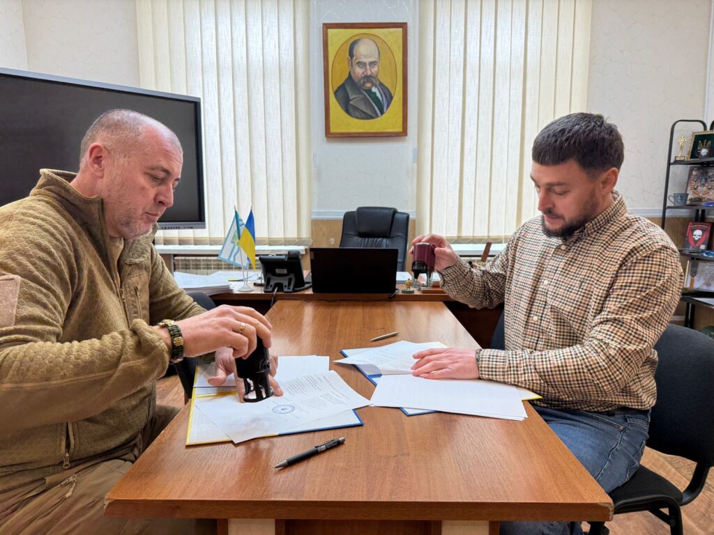 Двоє чоловіків сидять за столом, ставлять печатки на документи. Один — у коричневій флісовій куртці, інший — у клітчастій сорочці. На столі — папери, ручка, папки. Позаду — монітор, телефон, два прапорці (український і ще один), портрет на стіні, полиця з предметами.