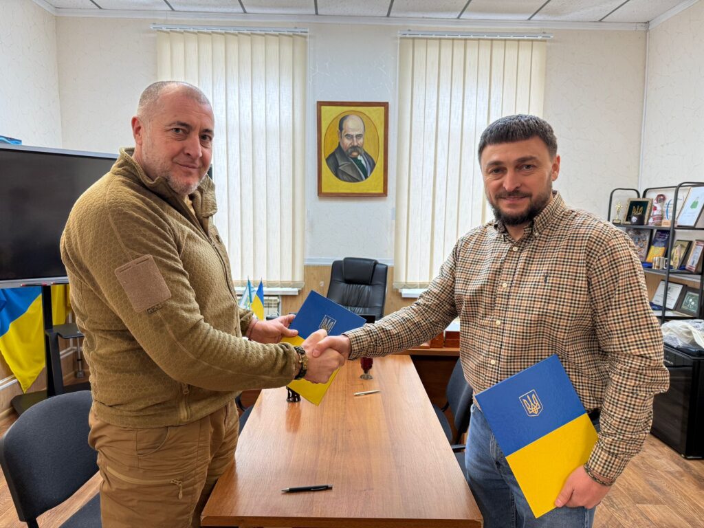 Двоє чоловіків стоять по обидва боки дерев’яного столу, потискають руки. В руках — синьо-жовті папки з гербом України. Позаду — великі вікна з жалюзі, портрет на стіні, українські прапори, монітор і полиця з нагородами.