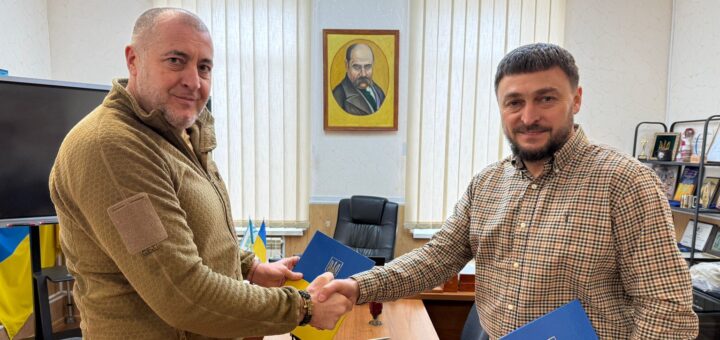 Двоє чоловіків стоять по обидва боки дерев’яного столу, потискають руки. В руках — синьо-жовті папки з гербом України. Позаду — великі вікна з жалюзі, портрет на стіні, українські прапори, монітор і полиця з нагородами.