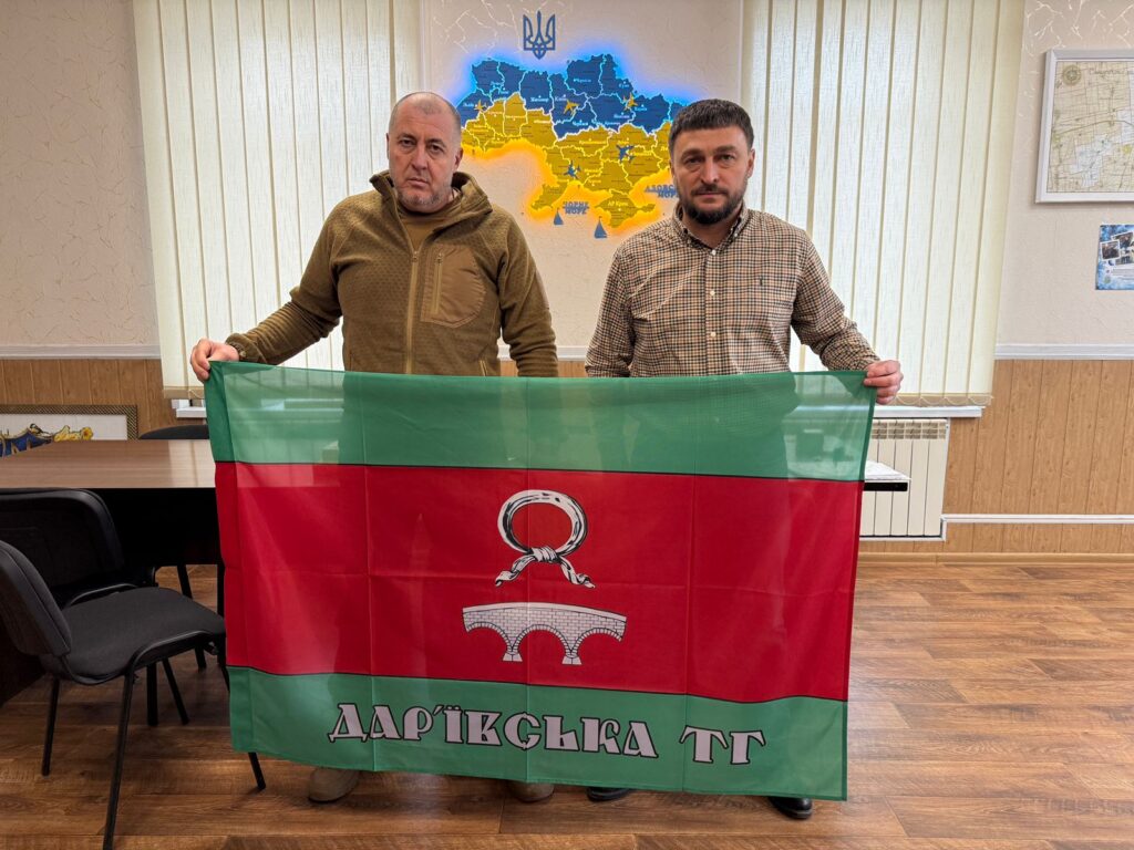 Двоє чоловіків тримають прапор із зеленими краями та червоним центром. У центрі — графіка кам’яного мосту з трьома арками й мотузковим кільцем, напис «ДАР'ЇВСЬКА ТГ». Позаду — карта України в синьо-жовтих кольорах і герб. 
