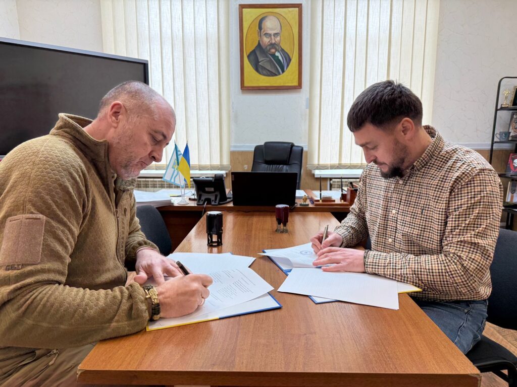 Двоє чоловіків сидять за дерев’яним столом, підписують документи. Один — у коричневому тактичному светрі, інший — у клітчастій сорочці