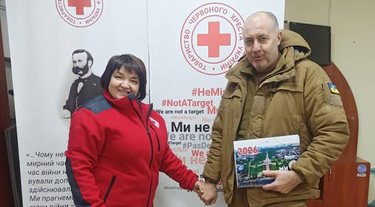 У приміщенні стоять двоє людей перед двома вертикальними банерами Червоного Хреста. Один — у червоно-чорній куртці з емблемою організації, другий — у військовій формі, тримає календар із написом «2026». Вони тримаються за руки й усміхаються. Банери містять емблеми, цитати українською про захист мирного населення, заклики до поваги міжнародного гуманітарного права та хештеги: «#HeMиЦіль», «#NotATarget».