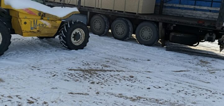 На засніженій території телескопічний навантажувач New Holland LM410 вивантажує великі прямокутні коробки з напівпричепа вантажівки. Бічна завіса причепа відкрита, видно синій внутрішній простір і штабелі коробок. На землі — сліди шин, сонце світить яскраво.