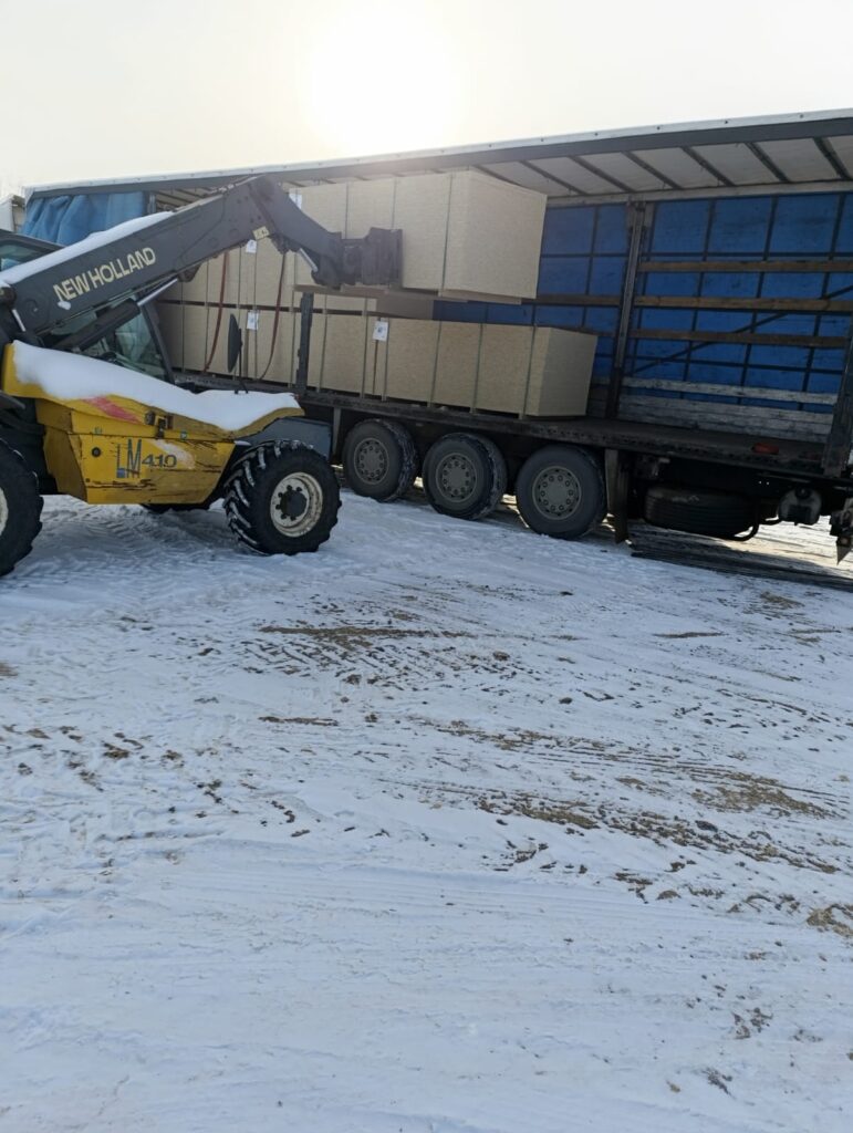 На засніженій території телескопічний навантажувач New Holland LM410 вивантажує великі прямокутні коробки з напівпричепа вантажівки. Бічна завіса причепа відкрита, видно синій внутрішній простір і штабелі коробок. На землі — сліди шин, сонце світить яскраво.