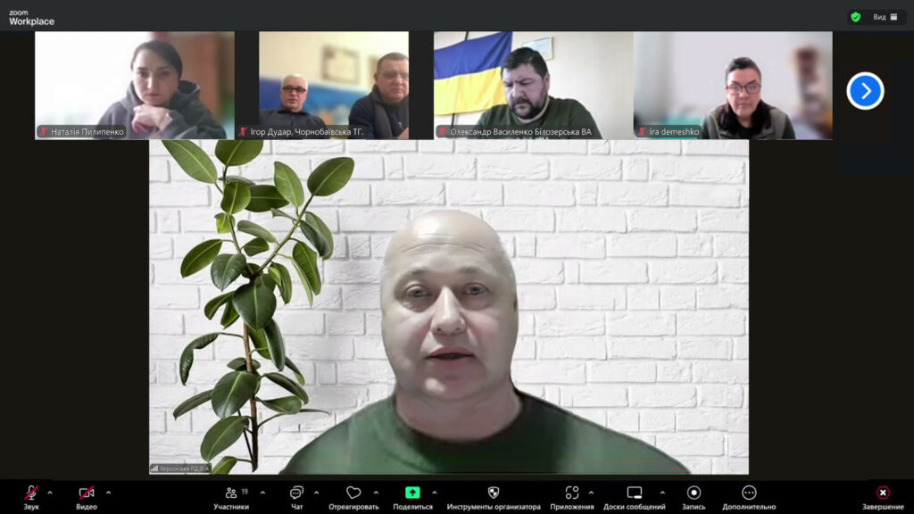 Скріншот Zoom-конференції. Головний спікер —Жадан Михайло Анатолійович, у центрі, на фоні білої цегляної стіни з зеленою рослиною. У верхньому ряду — п’ять менших відео інших учасників. Інтерфейс — Zoom Workplace, з кнопками керування внизу: звук, відео, чат, учасники, запис, завершення тощо.