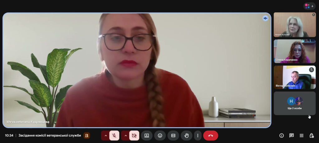 Скріншот онлайн-зустрічі, головна учасниця — Кудрявцева, у червоному светрі, з довгою косою та окулярами. Позаду — зелена рослина, біла ваза, книжки на тумбі. Праворуч — мініатюрні вікна інших учасників: Наталя Устінова, Олена Кириченко, Віктор Колісник, ще двоє позначені як «Ще 2 особи». Час — 10:34, назва зустрічі — «Засідання комісії ветеранської служби». 