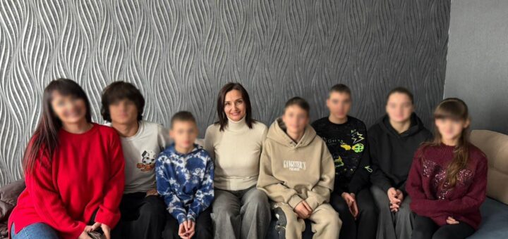 Вісім людей сидять на дивані біля стіни з хвилястим сірим візерунком. Одяг — повсякденний, обличчя розмиті для конфіденційності.