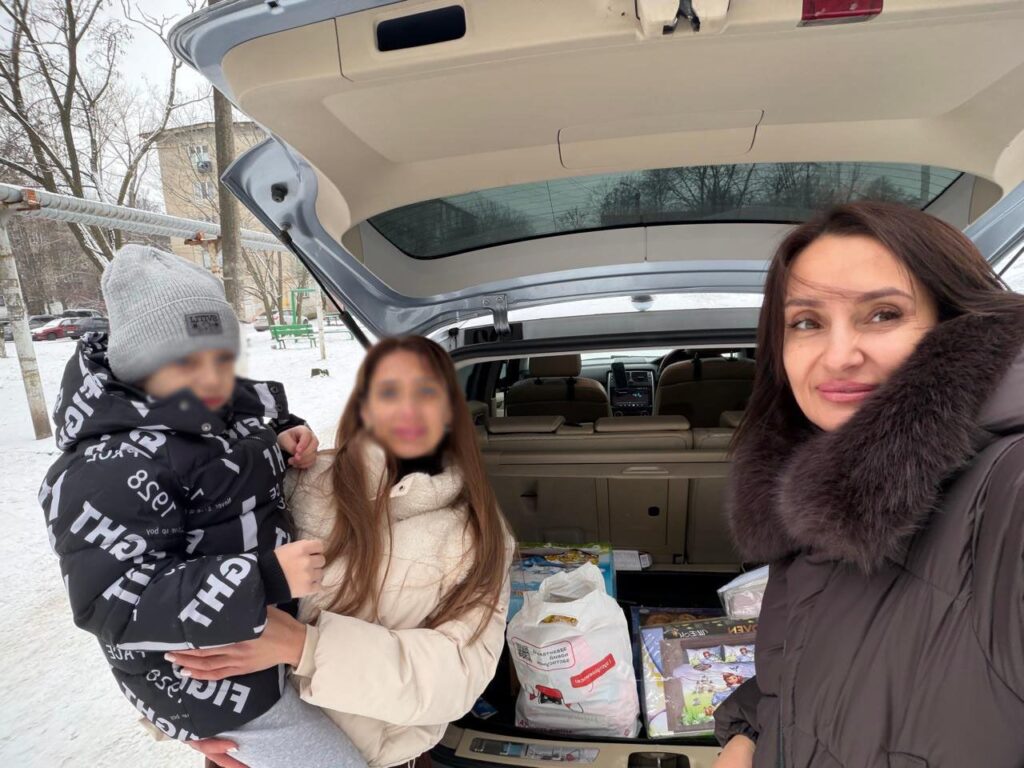 На вулиці, серед снігу, троє людей біля відкритого багажника автомобіля. Один дорослий тримає дитину в чорній куртці з білим написом і сірій шапці. Інший дорослий — у темному пальті з хутряним капюшоном. У багажнику — пакети, зокрема з написами «Пятёрочка» і «Детский мир». Позаду — дерева без листя й житловий будинок.