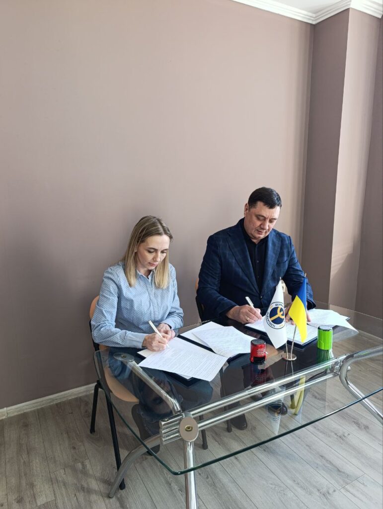 Двоє людей сидять за скляним столом і підписують документи. На столі — папери, ручки, червона печатка, зелений контейнер і два прапорці