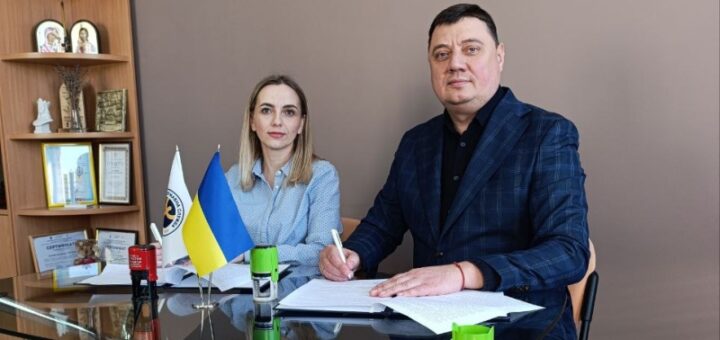 Чоловік у синьому костюмі в клітинку сидить праворуч і підписує документ. Ліворуч — жінка у світло-блакитній сорочці. На столі — документи, зелена чашка, банка Coca-Cola, два маленькі прапорці (український та інший з емблемою). Позаду — дерев’яна полиця з грамотами, іконами та декоративними речами. А