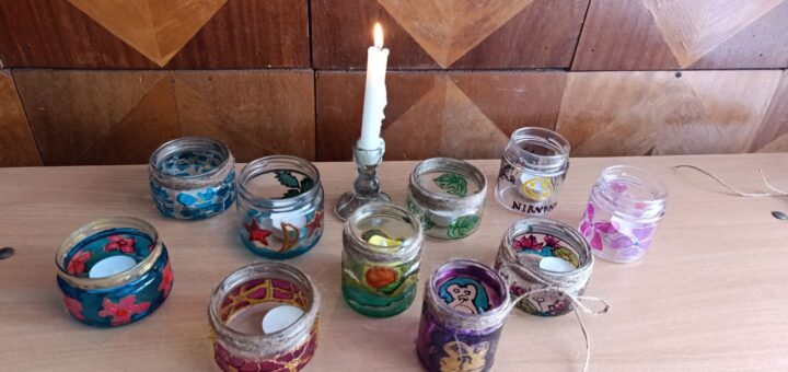 На дерев’яній поверхні перед стіною з геометричним дерев’яним оздобленням розташовано десять маленьких скляних баночок. Кожна прикрашена яскравими орнаментами: квітковими, абстрактними, мультяшними. Деякі обмотані мотузкою, кілька містять маленькі свічки. У центрі — біла свічка в металевому тримачі, запалена.