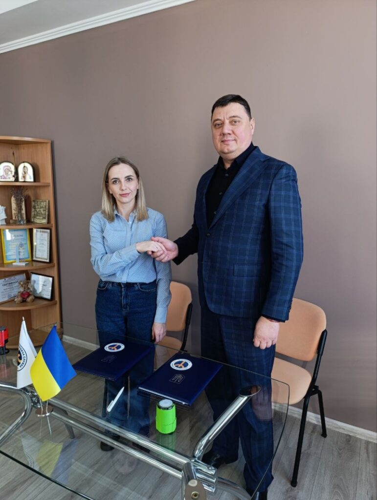 Двоє людей стоять і потискають руки через скляний стіл. Чоловік — у темно-синьому костюмі в клітинку, жінка — у світло-блакитній сорочці та джинсах. На столі — дві сині папки з емблемою, зелена чашка, ручка та два маленькі прапорці: України й інший з логотипом. Позаду — дерев’яна полиця з грамотами, нагородами та іконами.