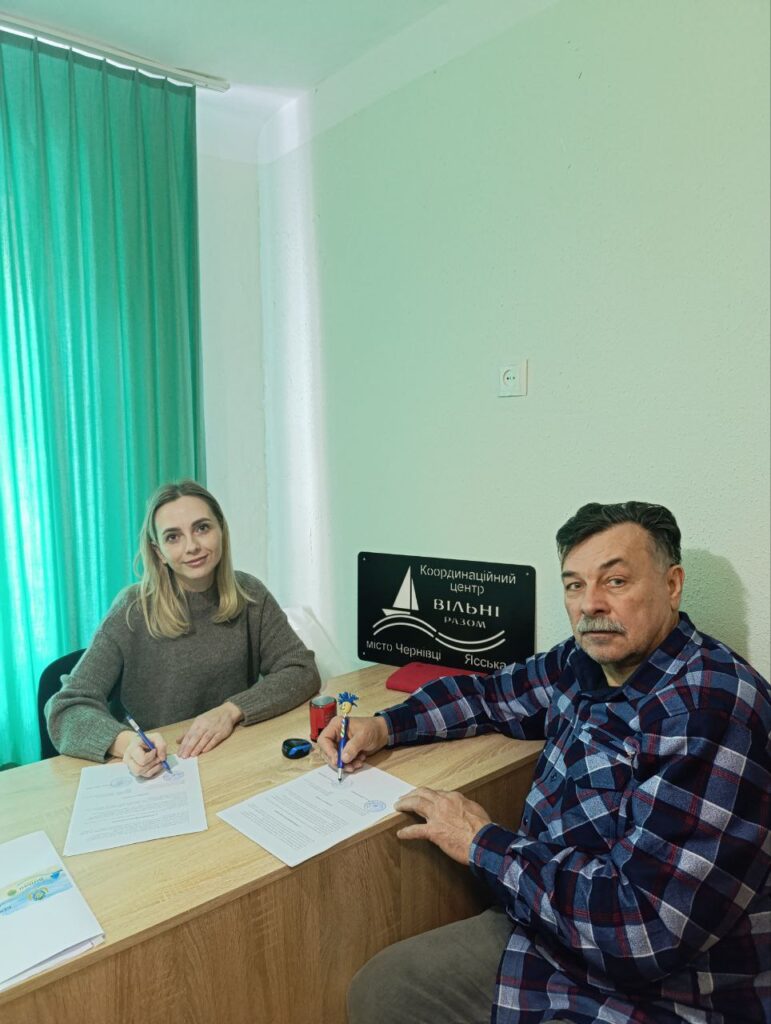За дерев’яним столом сидять двоє людей — жінка в сірому светрі та чоловік у клітчастій сорочці. Обоє тримають ручки й підписують документи. На столі — кілька паперів, червона чашка, окуляри та ручка з декоративним елементом. На стіні позаду — чорна табличка з білим написом: «Координаційний центр Вільні разом місто Чернівці Ясська». 