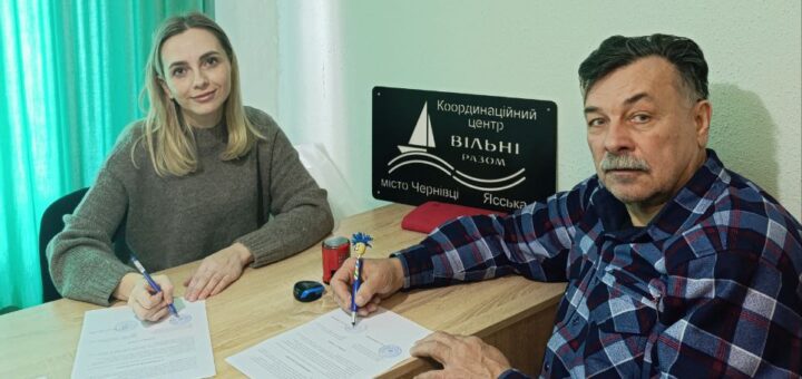 За дерев’яним столом сидять двоє людей — жінка в сірому светрі та чоловік у клітчастій сорочці. Обоє тримають ручки й підписують документи. На столі — кілька паперів, червона чашка, окуляри та ручка з декоративним елементом. На стіні позаду — чорна табличка з білим написом: «Координаційний центр Вільні разом місто Чернівці Ясська».