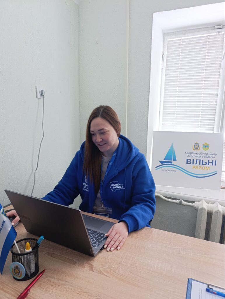 Людина сидить за столом із ноутбуком, одягнена в синю худі з написами «Право на захист» та «UNHCR». На столі — ручки, прапорець. Праворуч — табличка з написом «Координаційний центр Херсонська область Вільні разом місто Чернівці» та логотипи організацій. 