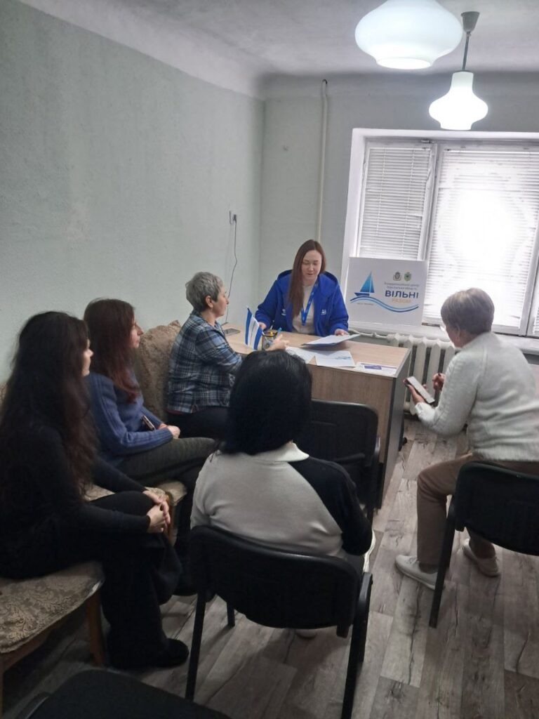 У кімнаті з зеленими стінами та дерев’яною підлогою шестеро людей. П’ятеро сидять, один стоїть за столом у синій куртці з бейджем. На столі — папери та маленькі прапорці. Позаду — табличка з написом «Вільні разом» і логотип із вітрильником. 