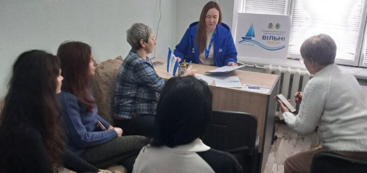 У кімнаті з зеленими стінами та дерев’яною підлогою шестеро людей. П’ятеро сидять, один стоїть за столом у синій куртці з бейджем. На столі — папери та маленькі прапорці. Позаду — табличка з написом «Вільні разом» і логотип із вітрильником.