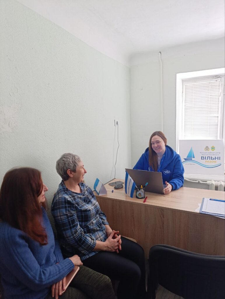 Невеликий офіс. За столом сидить чоловік у синій худі з білим написом, перед ним ноутбук, ручки та документи. Двоє людей навпроти уважно слухають. На столі — таблички з іменами. Позаду — табличка з написом «Координаційний центр підтримки цивільного населення Вільні разом» та логотип із вітрильником