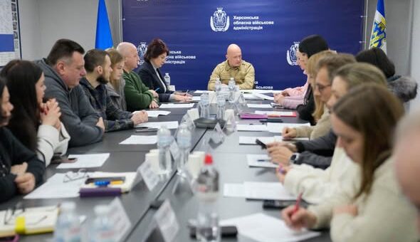 У просторій залі за довгим столом сидять численні учасники, ведуть обговорення та роблять нотатки. На столі — пляшки з водою, документи, таблички з іменами. Позаду — синій банер із білим написом «Херсонська обласна військова адміністрація» та державні прапори України з обох боків.