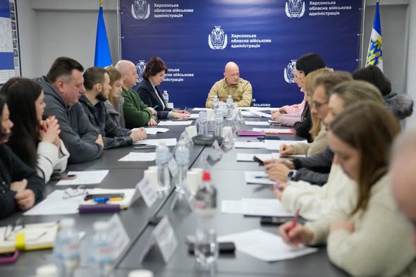 У просторій залі за довгим столом сидять численні учасники, ведуть обговорення та роблять нотатки. На столі — пляшки з водою, документи, таблички з іменами. Позаду — синій банер із білим написом «Херсонська обласна військова адміністрація» та державні прапори України з обох боків. 