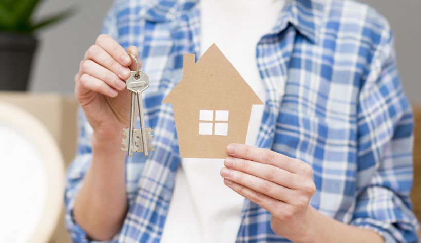 Людина в синій картатій сорочці тримає в руках два предмети: у правій — металеві ключі на брелоку, у лівій — картонний виріз у формі будинку з білим віконцем. 