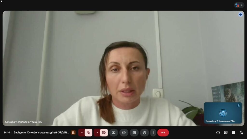 Скріншот відеоконференції. У центрі — жінка в білому светрі, сидить перед світлою стіною з розеткою та вертикальним кабелем. Позаду — рослина й частково видимий плакат або календар. У нижній панелі — кнопки керування: мікрофон, камера, демонстрація екрану, завершення дзвінка. Текст угорі: «Служба у справах дітей ХРВА». Атмосфера — офіційна, робоча.