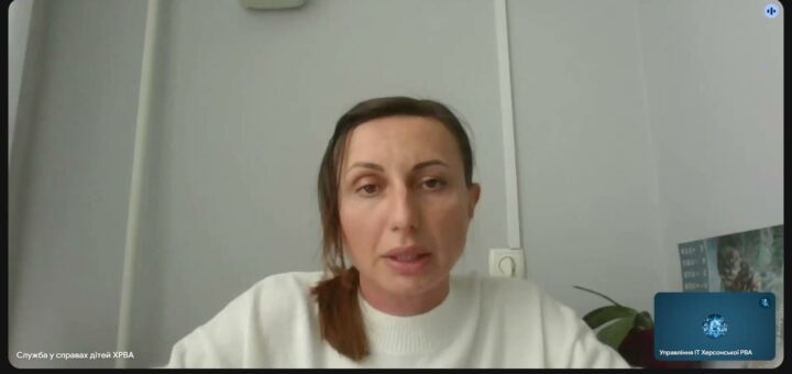 Скріншот відеоконференції. У центрі — жінка в білому светрі, сидить перед світлою стіною з розеткою та вертикальним кабелем. Позаду — рослина й частково видимий плакат або календар. У нижній панелі — кнопки керування: мікрофон, камера, демонстрація екрану, завершення дзвінка. Текст угорі: «Служба у справах дітей ХРВА». Атмосфера — офіційна, робоча.