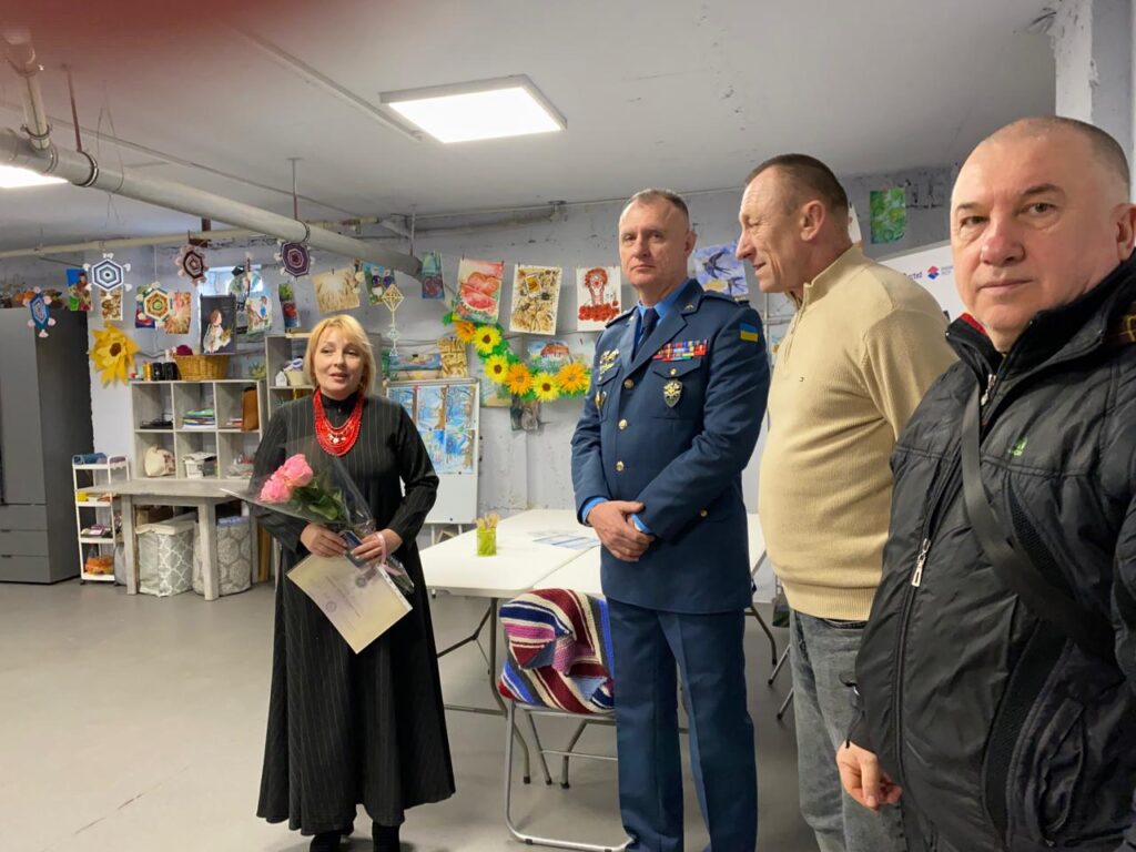 Чотири дорослі стоять у яскраво прикрашеній кімнаті. Жінка зліва тримає букет рожевих троянд і сертифікат, одягнена в довгу чорну сукню з червоним намистом. Поруч — чоловік у синій військовій формі з медалями та прапором України на рукаві. Двоє інших чоловіків — у повсякденному одязі (бежевий светр і чорна куртка). Кімната прикрашена паперовими соняхами, геометричними орнаментами, малюнками й виробами, на полицях — матеріали для творчості. 