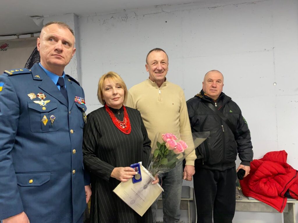 Чотири людини стоять біля стіни. Ліворуч — чоловік у синій військовій формі з медалями та шевроном із прапором України. Поруч — жінка в чорній сукні у смужку з червоним намистом, тримає букет рожевих троянд і сертифікат. Біля неї — чоловік у бежевому светрі та джинсах, усміхається. Праворуч — чоловік у чорній куртці