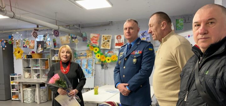 Чотири дорослі стоять у яскраво прикрашеній кімнаті. Жінка зліва тримає букет рожевих троянд і сертифікат, одягнена в довгу чорну сукню з червоним намистом. Поруч — чоловік у синій військовій формі з медалями та прапором України на рукаві. Двоє інших чоловіків — у повсякденному одязі (бежевий светр і чорна куртка). Кімната прикрашена паперовими соняхами, геометричними орнаментами, малюнками й виробами, на полицях — матеріали для творчості.