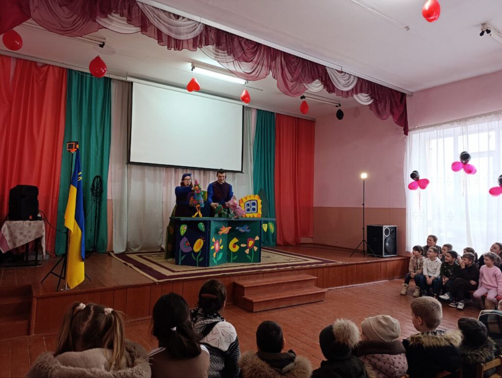 Двоє артистів за ширмою керують ляльками, перед ними сидять діти. Сцена прикрашена червоними, зеленими та білими завісами, над нею — червоні кульки. Ліворуч — прапор України.