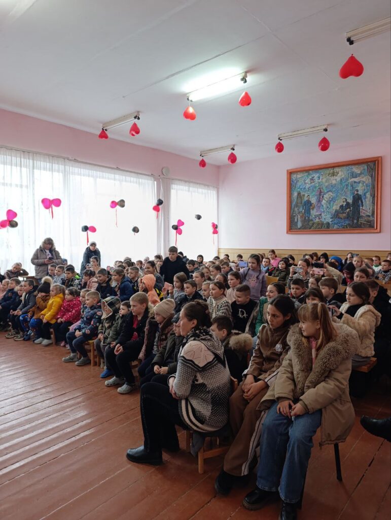 Велика група дітей і кілька дорослих сидять у кімнаті з рожевими стінами та дерев’яною підлогою. На стелі висять червоні й рожеві сердечка, що створюють святковий настрій. На стіні — велика картина з багатофігурною сценою.